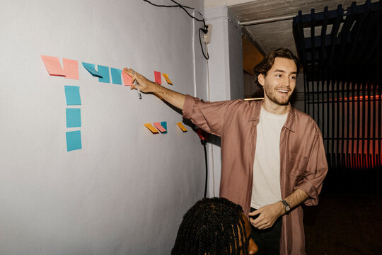 Smiling male entrepreneur sticking adhesive notes on wall in meeting at office