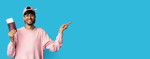 smiling indian man with plane tickets and passport posing over blue studio background, pointing at copy space, panorama. Stylish middle eastern hipster travelling while COVID-19 pandemic