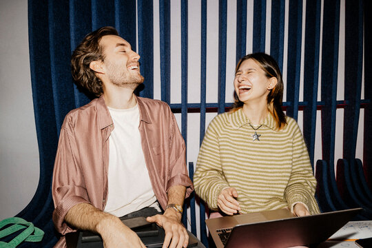 Happy male and female business colleagues laughing while sitting with laptops at office
