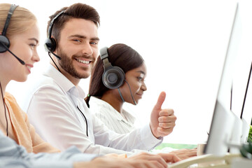 Happy male call centre operator showing thumb up gesture, working with his colleages at modern...