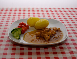 A savory dish features tender pieces of pork in rich sauce alongside two boiled potatoes and a fresh salad of cherry tomatoes and cucumber