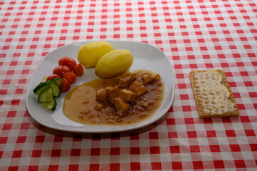A delicious plate showcases pork pieces in savory sauce accompanied by boiled potatoes, cherry tomatoes, and cucumber slices, all placed on a checkered tablecloth