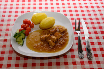 A satisfying plate features pork pieces in a rich sauce, served with boiled potatoes and a side of fresh cucumber and cherry tomatoes on a vibrant red and white checkered tablecloth
