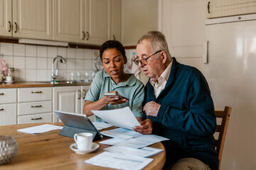 Female healthcare worker using smart phone while helping senior man in examining medical test results at home