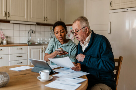 Female healthcare worker using smart phone while helping senior man in examining medical test results at home