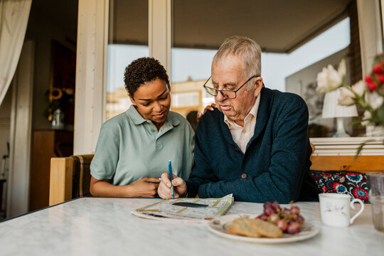 Senior man solving crossword puzzle in newspaper while sitting next to female nurse outside house