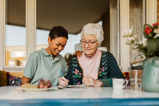 Senior woman solving crossword puzzle in newspaper while sitting next to female nurse outside house