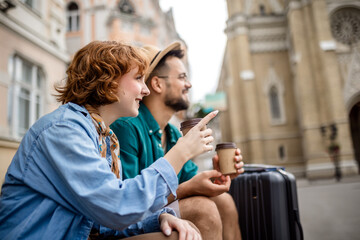 Ginger girlfriend and her boyfriend sitting in the city center and drink coffee. They are on vacation and have their luggages and backpacks with them.