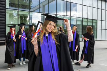Portrait of graduate student standing in front of her friends with toga and diploma all happy.