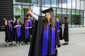 Portrait of graduate student standing in front of her friends with toga and diploma all happy.