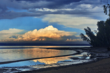 A picturesque landscape, with a thundercloud hanging in the sky above the river, illuminated by the sunlight.