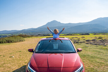 Little girl joyfully sticks out of car sunroof with raised hands against the backdrop of mountains...