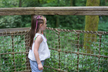 Naklejka premium A little girl with small multicolored braids on a hanging rope bridge in a summer park. Concept of children's activity and leisure.