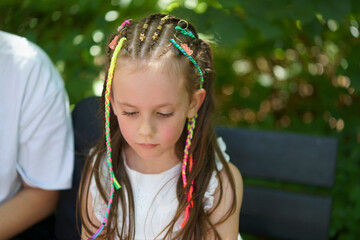 A sad little girl with small multicolored braids sits on a bench in a summer park. Concept of...