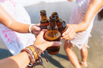 Close-up of several hands clinking beer bottles together in a toast on a sunny beach, symbolizing fun, friendship, and celebration against the backdrop of ocean waves and blue skies.