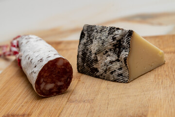 a wedge of cured sheep's cheese with a moldy rind and a piece of salami with its cord on a wooden cutting board on a white-veined marble table