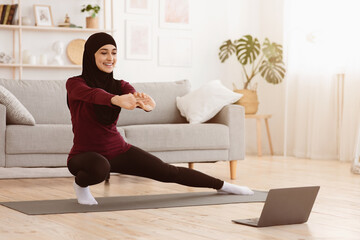 Happy arabic woman in hijab doing lunges in front of laptop at home, making fitness training via online tutorials, copy space