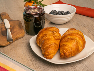 Croissants, jar of jam, butter knife, wooden cutting board and fresh blueberries on wooden table.
