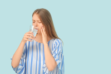 Young woman with sensitive teeth drinking cold water on blue background