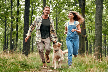Obraz premium A Joyful Couple Happily Running with Their Dog in a Lush and Green Forest Setting