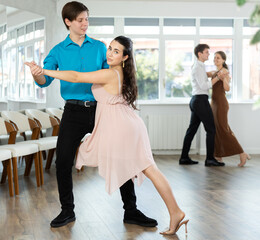 Smiling young brunette in light flowing dress performing elegant tango with stylish male partner in...
