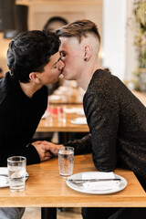 A vertical close up of a gay men couple on a date in a cozy cafe
A close up of a LGBTQ+ couple on a romantic date, a man timidly touching his partner's hand and kissing him across a table in an urban 