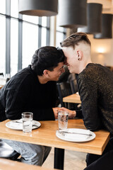 A vertical medium shot of a gay men couple on a date in a cozy cafe
A mid shot of a LGBTQ+ couple on a romantic date, a man timidly touching his partner's hand and laughing in an urban cafe
