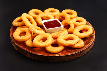 Wooden board with fried breaded onion rings and ketchup on black background