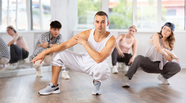 Young teacher and students perform hip hop dance. Squatting down, students make basic movements to dynamic music