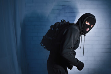 Burglary Concept. Sneaky and scared intruder wearing black balaclava hat lurking in the dark,...