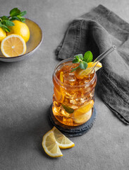 Iced tea with lemon, ice and mint in a glass on gark gray background with citrus fruits and napkin.