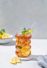 Iced tea with lemon, ice and mint in a glass on light texture background with citrus fruits and shadow.