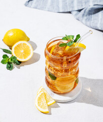 Iced tea with lemon, ice and mint in a glass on light  background with shadow, citrus fruits and napkin.