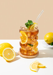 Iced tea with lemon, ice and mint in a glass on a white and beige background with shadow and citrus...