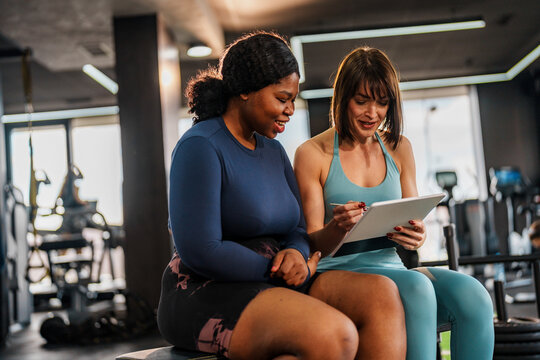 Smiling personal trainer presenting a training plan on a digital tablet to a cheerful overweight woman sitting in a gym, fostering motivation and support for her fitness journey