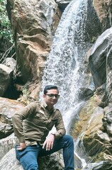 A man posing confidently in front of a stunning waterfall, surrounded by rugged rocks and lush greenery. The cascading water adds a serene and adventurous touch, capturing the beauty of nature.