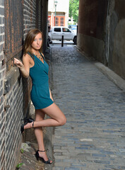 Beautiful young Caucasian female model poses in cobblestoned alley wearing a blue-green romper, standing next to brick wall