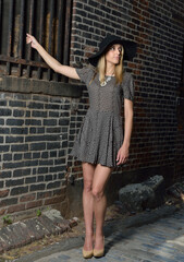 Beautiful young Hispanic female model poses in cobblestoned alley wearing a dark patterned dress and wide brimmed black hat
