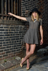 Beautiful young Hispanic female model poses in cobblestoned alley wearing a dark patterned dress and wide brimmed black hat