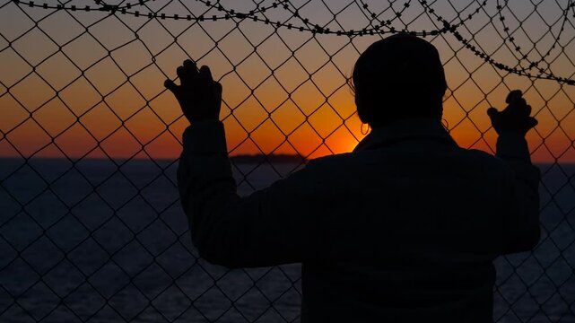 Refugee looking at sunset behind barbed wire fence. Silhouetted refugee gazing wistfully through barbed wire fence, golden sunset illuminating deep longing for freedom and hopes of future sanctuary