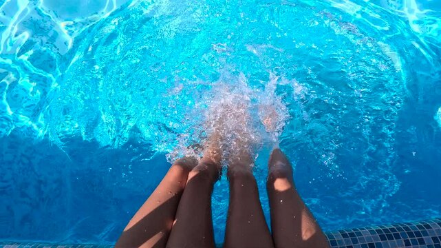 Fun in the outdoor pool. Two girls sit at the edge of a swimming pool, dangling their legs in the water and creating playful splashes, enjoying a sunny day filled with relaxation and fun