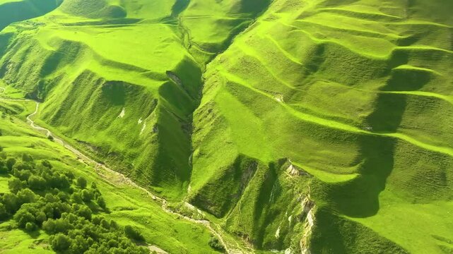 Drone shot of Maltas Canyon, Dagestan, with sunlight illuminating alpine-covered mountains, a flowing river, and dramatic cloud shadows over the landscape.