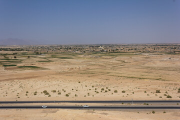 Scenic desert landscape near Abarkouh, Iran, with a highway, scattered shrubs, and distant farmlands under a clear blue sky.