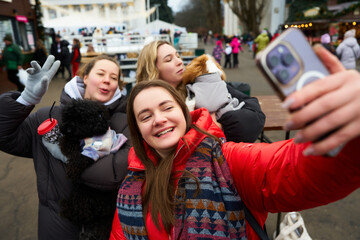 Friends take a joyful selfie with pets at a winter festival in a lively outdoor setting