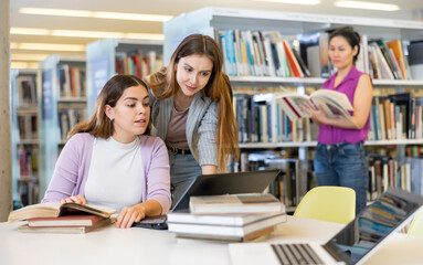 Two young women preparing to exam in library together, working on laptop computer, reading books...