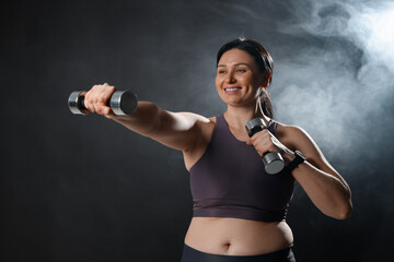 Plus size woman in gym clothes holding dumbbells on dark background with smoke