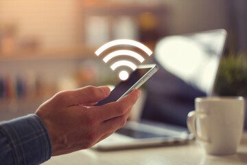 Man using a smartphone with a glowing wifi signal indoors near a laptop and a cup of coffee