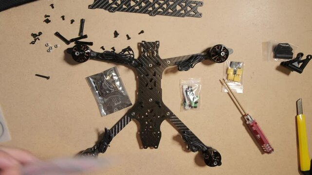 An engineer assembles a racing drone, carefully handling a flight controller. The drone's carbon fiber frame, brushless motors, and electronic components are laid out on a workbench, along with tools 