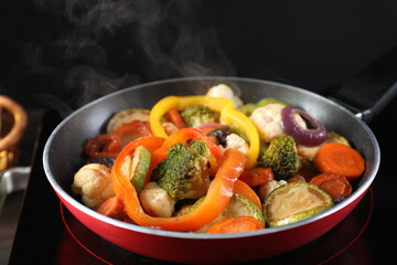 Frying pan with vegetables and stove on table against black background, closeup