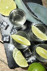 Tequila shots with lime slices, salt, ice cubes and agave leaves on wooden table, flat lay